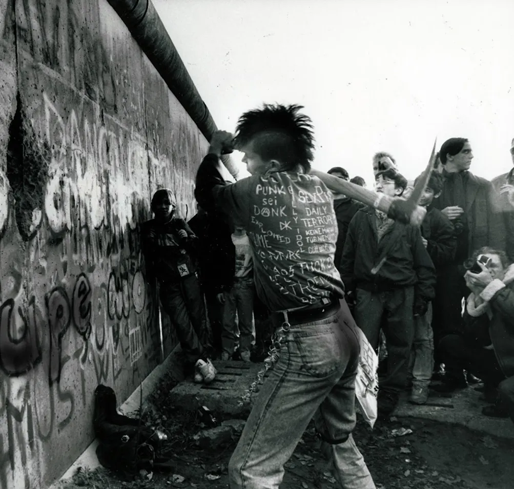A punk swinging a pickaxe at the graffiti-covered Berlin Wall while a crowd watches