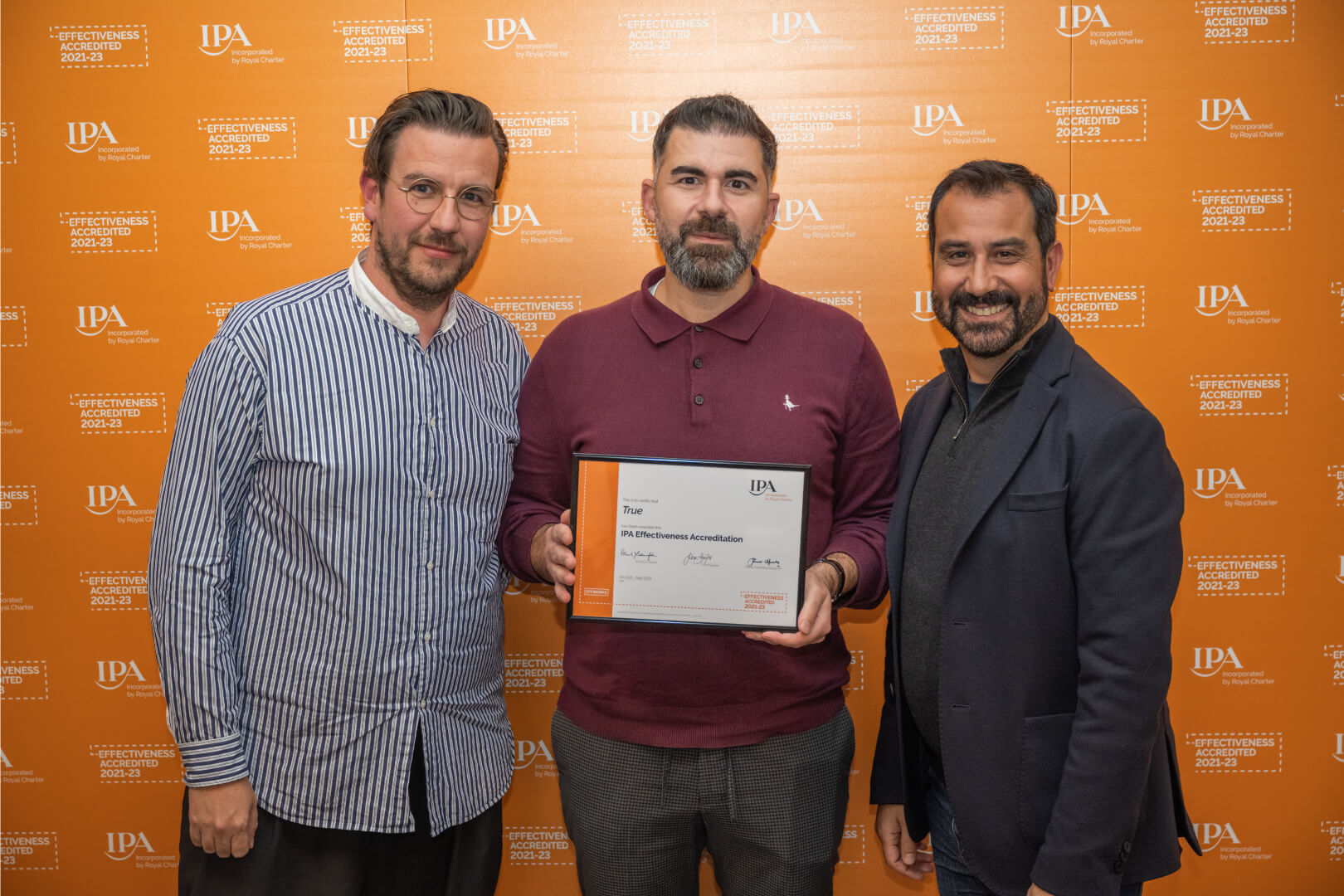 True's Founders & Head of Client Services standing in front of an orange background with the IPA logo on holding their IPA Effectiveness Accreditation certificate